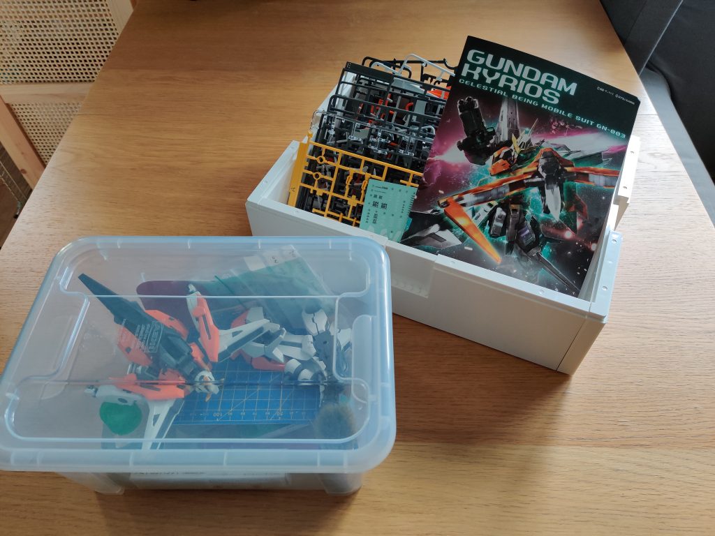 Transparent box containing a cutting mat and pieces of a plastic figurine; open-topped white box containing plastic sprew sheets and a "Gundam Kyrious" assembly manual.